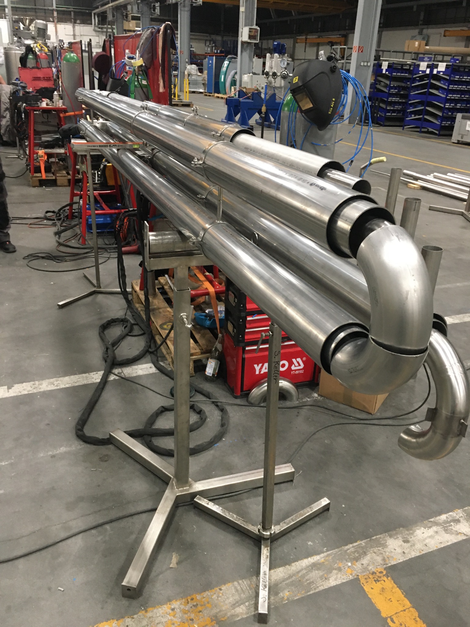Konstrukcja z polerowanych rur nierdzewnych na metalowym stojaku, z widocznymi spawami i zagięciami, w tle warsztat z narzędziami i osłoną spawalniczą.