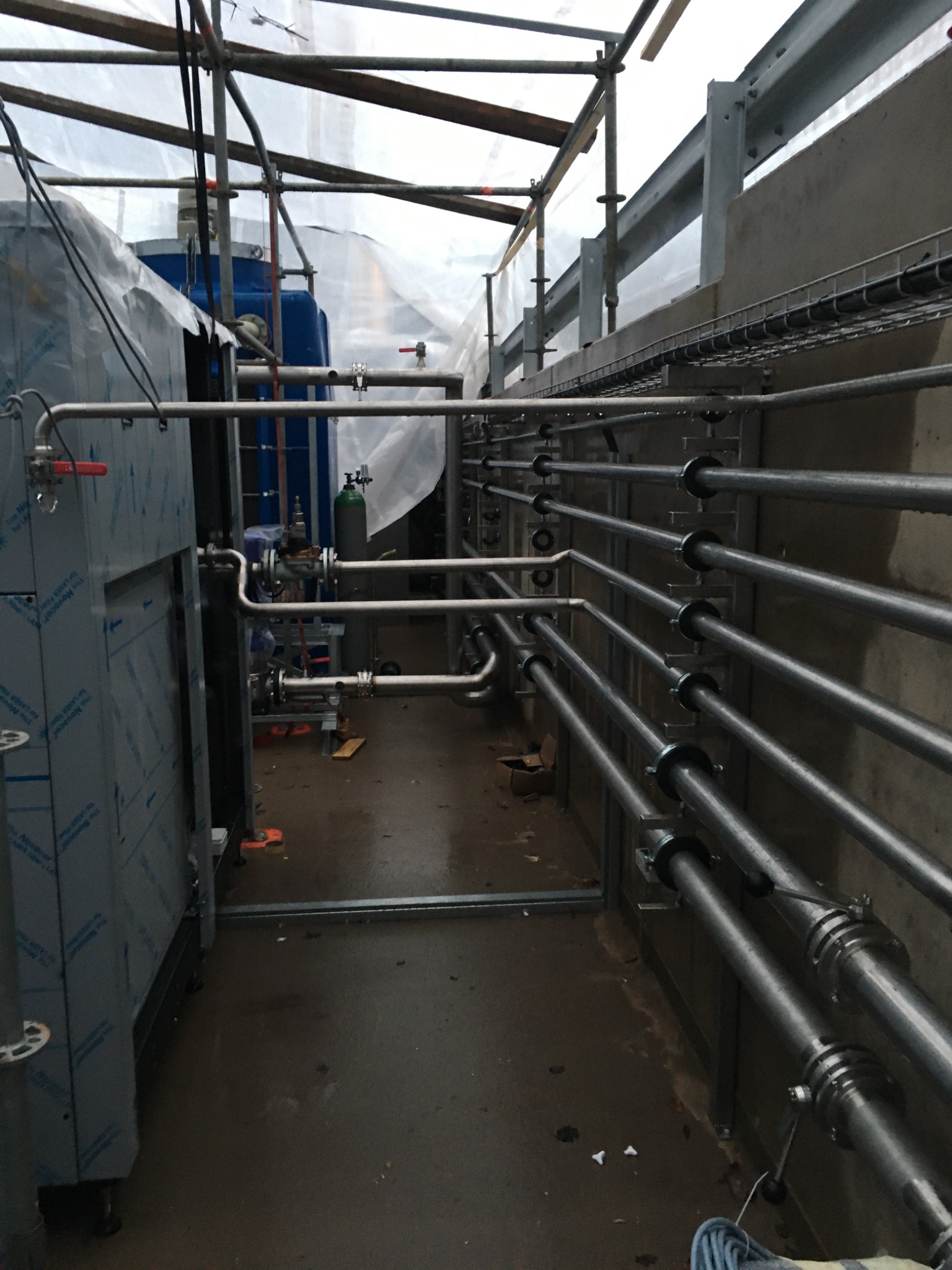 Instalacja przemysłowa z rurami ze stali nierdzewnej, widoczne elementy konstrukcyjne i zabezpieczenia budowlane, mokra posadzka.