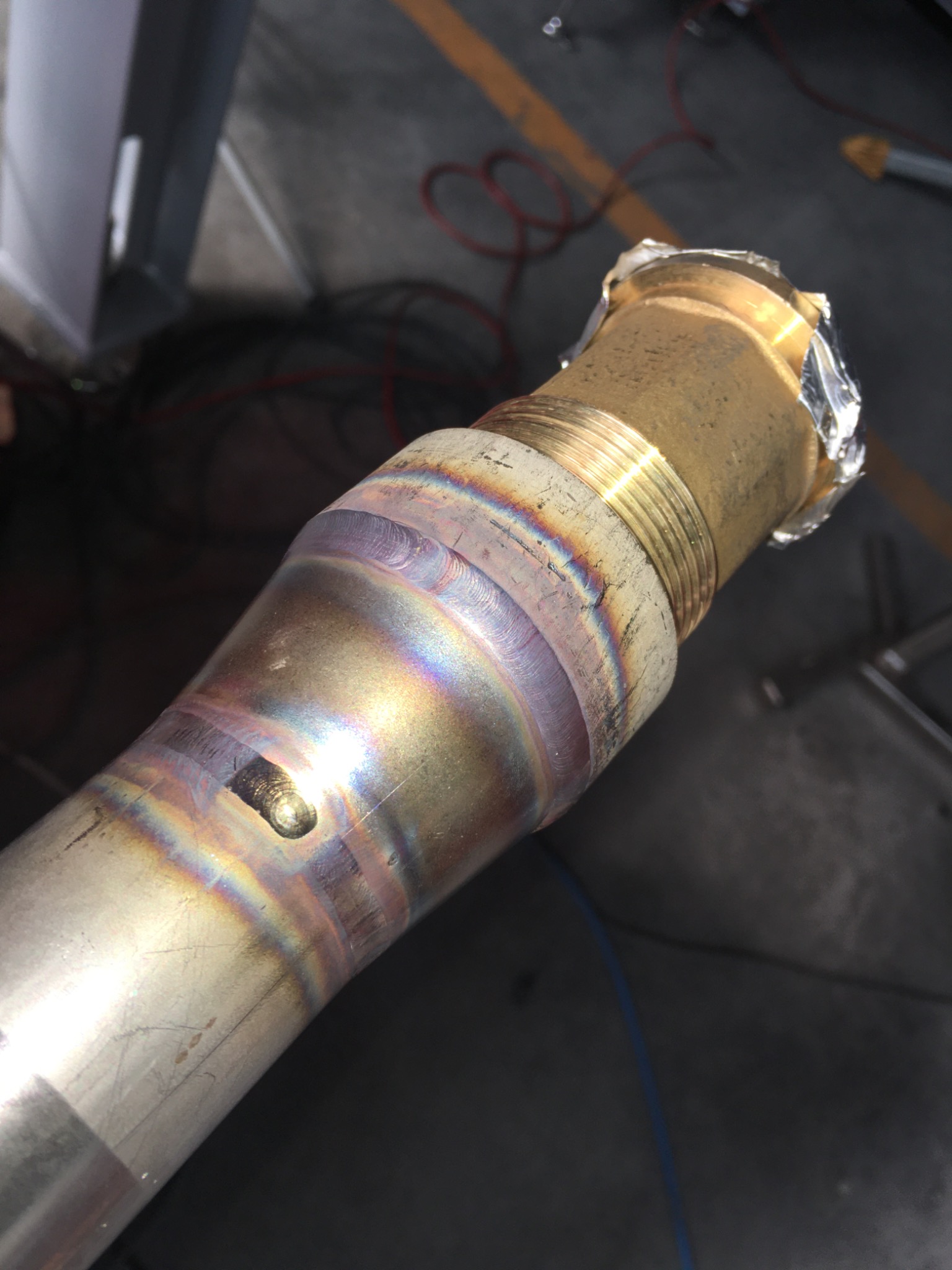 Metalowa rura z widocznymi śladami spawania, tęczowe zabarwienie metalu w miejscu łączenia, gwintowany element zabezpieczony folią.