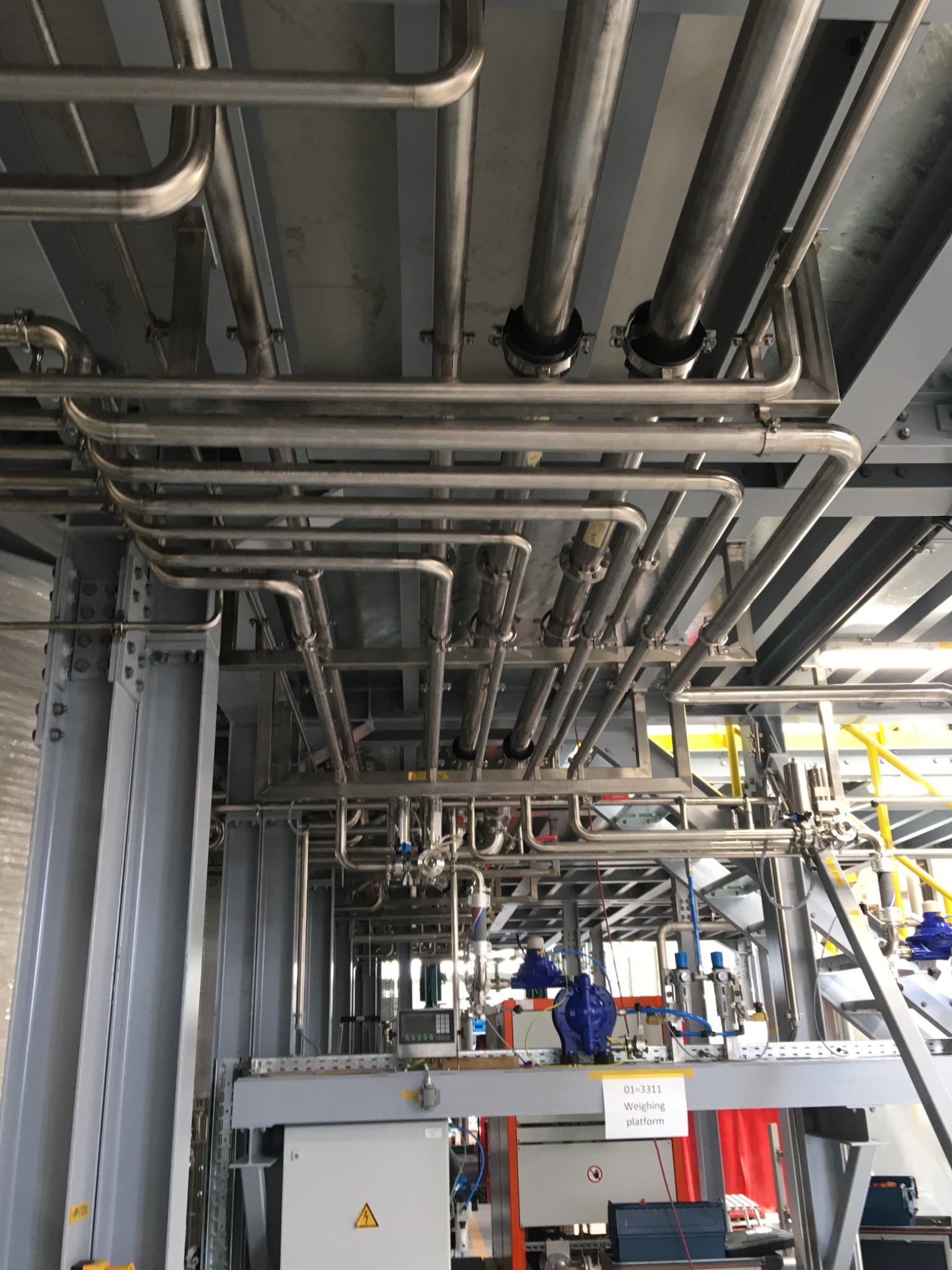 Skomplikowany układ rur ze stali nierdzewnej, zamocowanych pod sufitem konstrukcji stalowej, z widocznymi elementami instalacji technologicznej i tabliczką informacyjną.