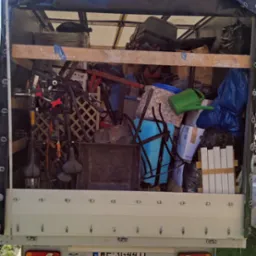 Wnętrze busa transportowego wypełnione r&oacute;żnorodnymi przedmiotami, w tym rowerami, meblami i kartonami, zabezpieczone czarną plandeką, widok z tyłu na tablicę rejestracyjną WE 3G4411.