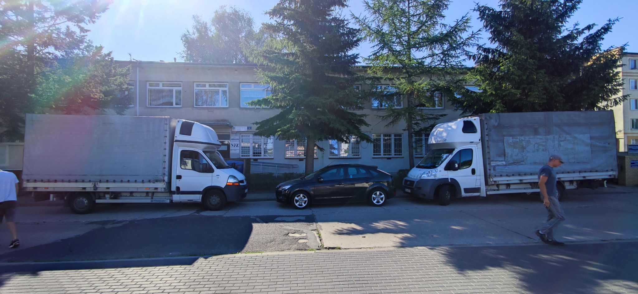 Dwa białe busy dostawcze z plandekami zaparkowane na chodniku przed budynkiem, obok stoi ciemny samochód osobowy, a obok jednego z busów przechodzi mężczyzna.