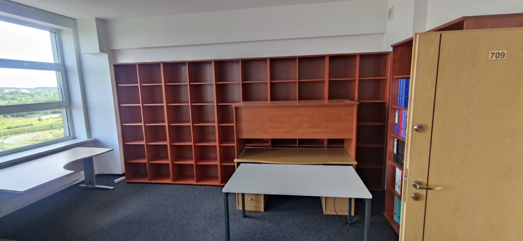 Pusta przestrzeń biurowa z regałami na ścianie, biurkami i szafą z numerem 709 na drzwiach.