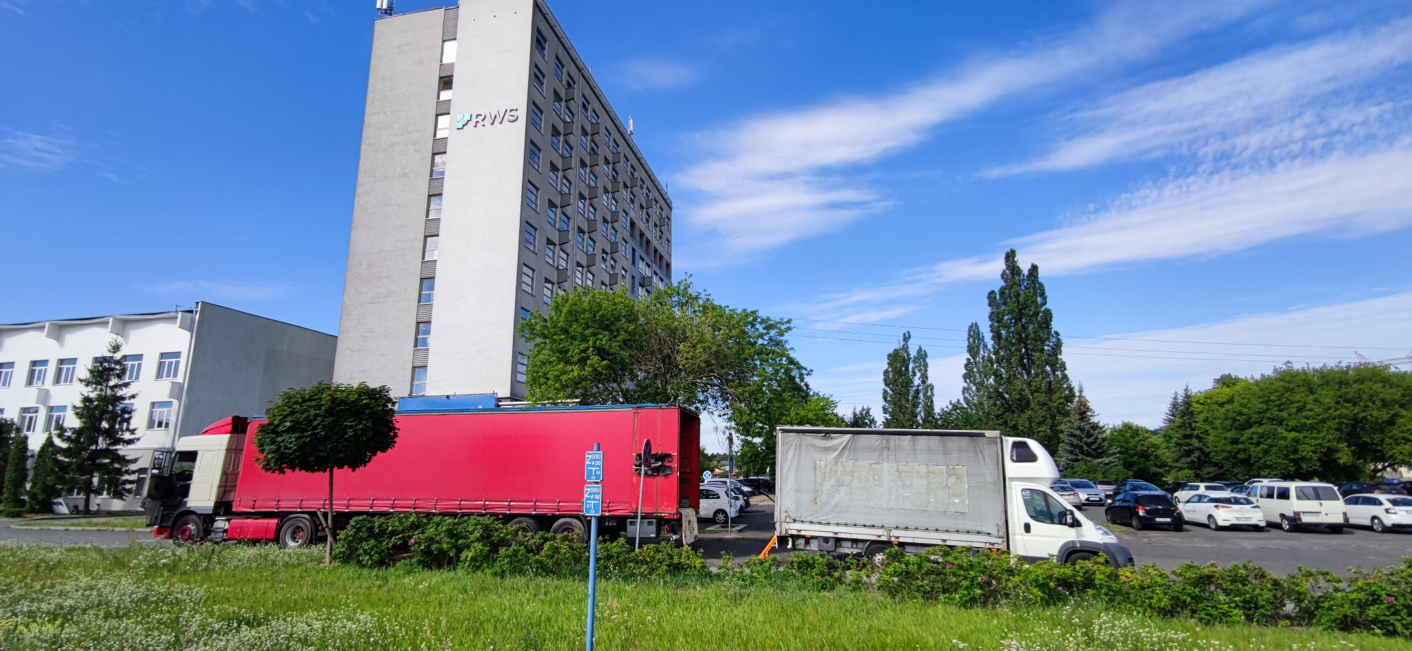 Dwa busy transportowe, czerwony i biały, zaparkowane na parkingu przed budynkiem RWS w słoneczny dzień.
