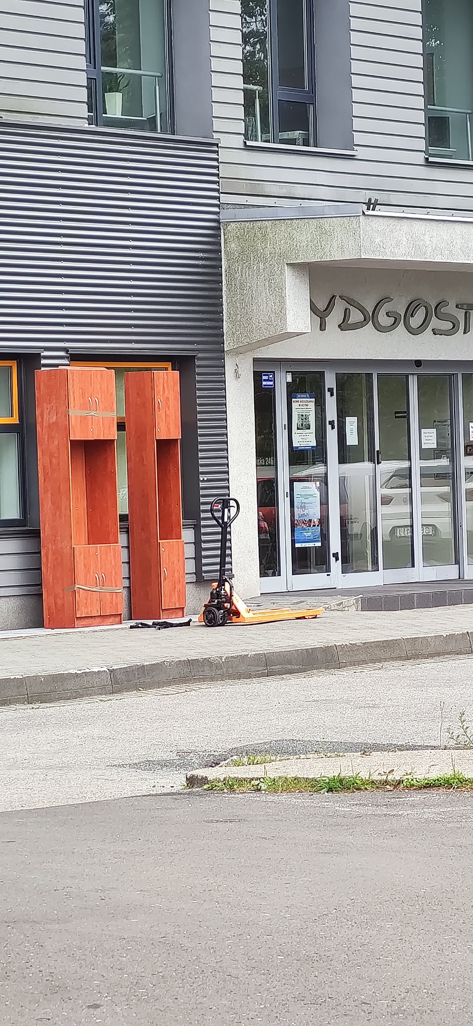 Dwa rozmontowane, drewniane segmenty szafy zabezpieczone taśmą stoją na chodniku przed budynkiem, obok wózek paletowy w kolorze pomarańczowym. Widoczne drzwi wejściowe do budynku z napisem 'BYDGOST'