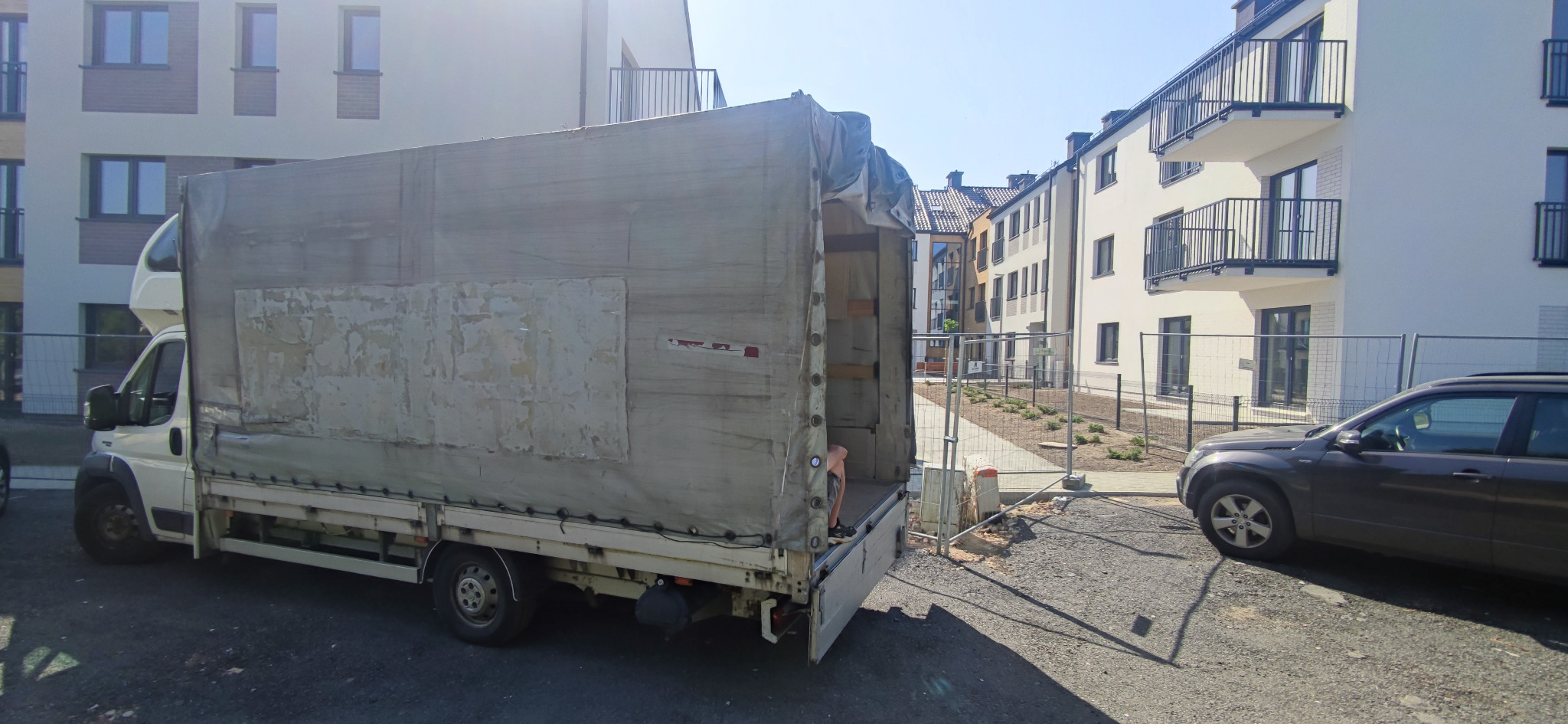 Biała furgonetka z plandeką na tle nowego osiedla mieszkaniowego, widoczne ślady użytkowania na plandece i otwarte drzwi przestrzeni ładunkowej.