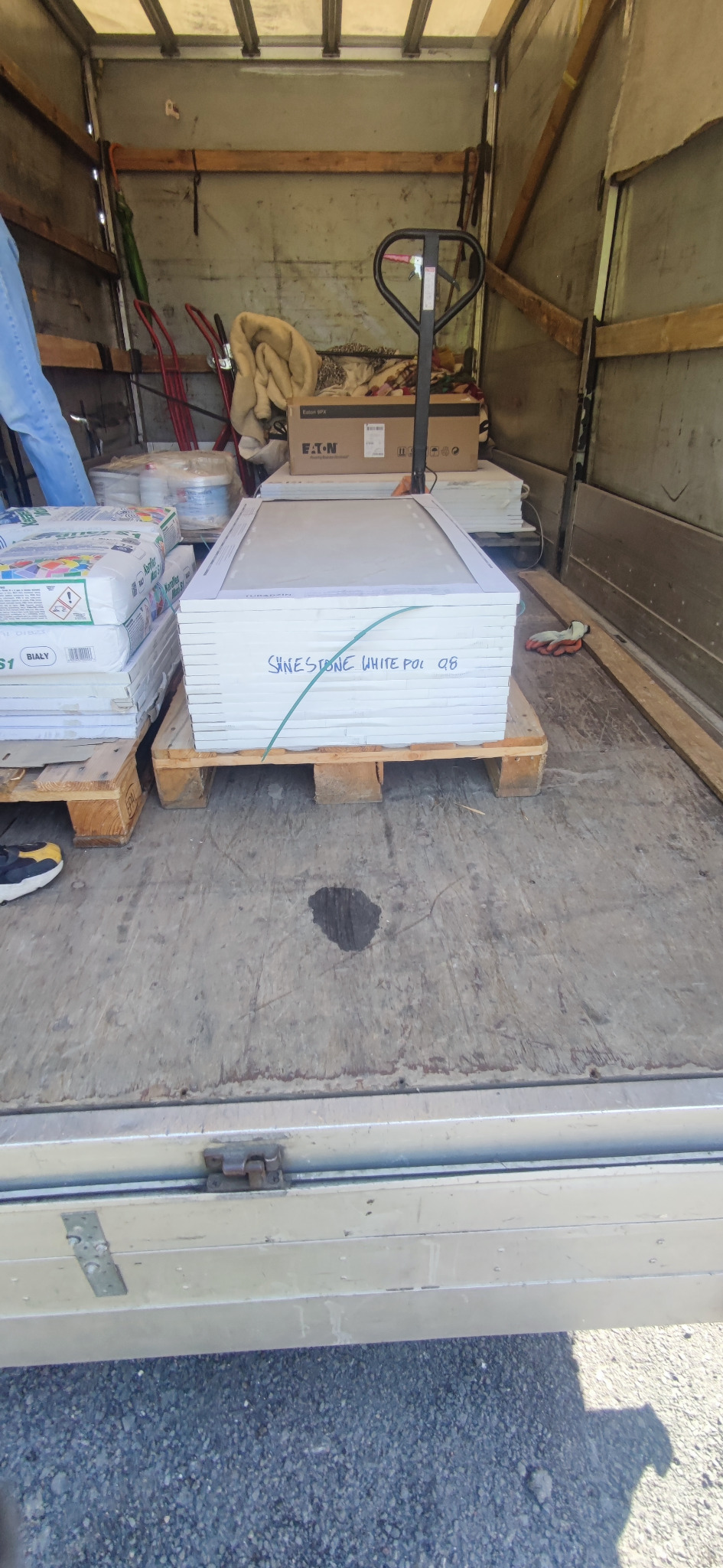 Palety z materiałami budowlanymi, w tym worki z zaprawą i płyty 'Sinestone White Pol Q8', załadowane do wnętrza samochodu dostawczego z widocznym wózkiem paletowym.