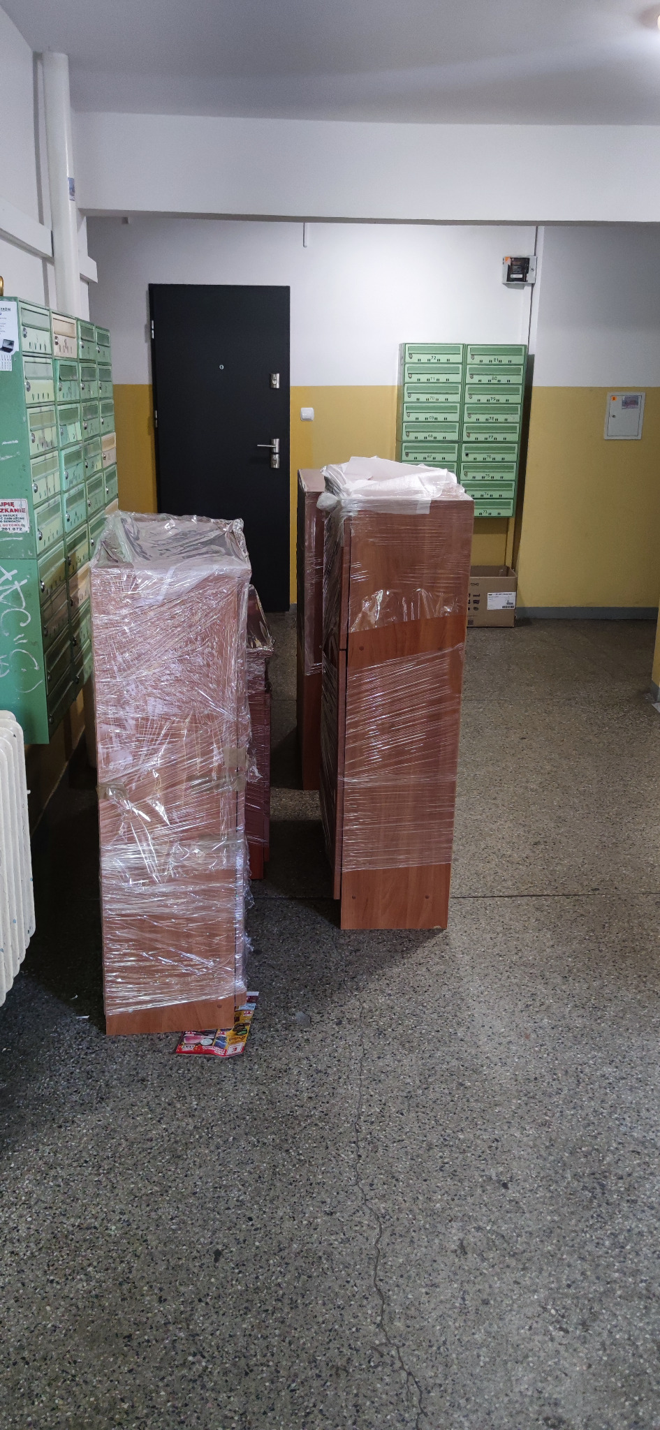Meble biurowe owinięte folią stretch, przygotowane do transportu, stoją w korytarzu bloku mieszkalnego, widoczne skrzynki pocztowe.