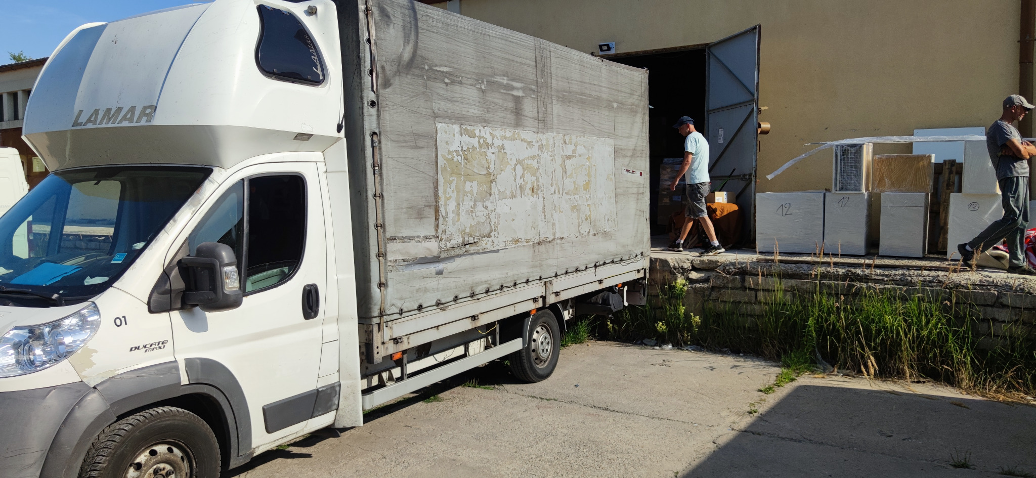 Biały samochód dostawczy marki Fiat Ducato z plandeką, zaparkowany przy magazynie podczas rozładunku towarów. Widoczni dwaj mężczyźni, jeden wchodzi do magazynu, drugi stoi obok zapakowanych paczek...