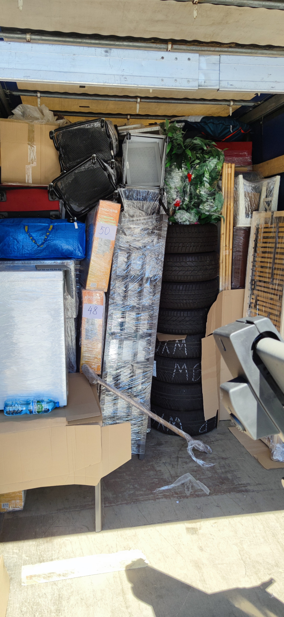 Wnętrze załadowanego busa: meble, kartony, opony samochodowe z białymi napisami, zapakowane rośliny i inne przedmioty zabezpieczone folią stretch.