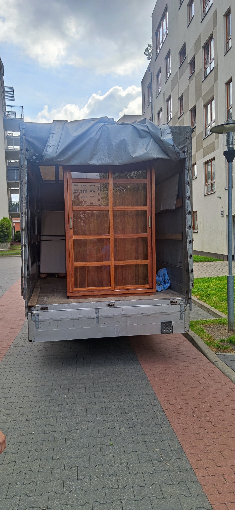 Drewniana szafa z przesuwanymi drzwiami zabezpieczona w przestrzeni ładunkowej samochodu dostawczego podczas transportu, widok z zewnątrz na tle budynków mieszkalnych.