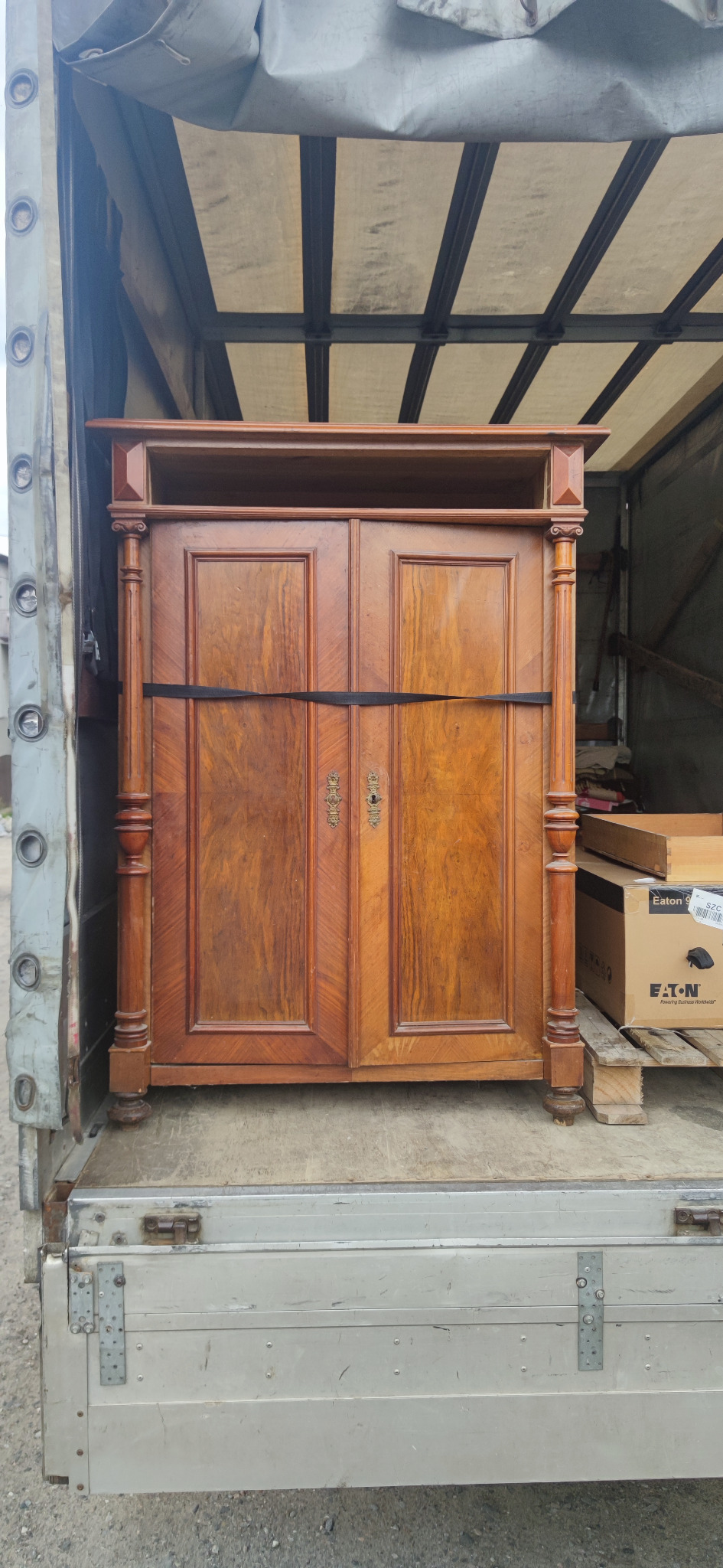 Antyczny, drewniany kredens zabezpieczony czarnym pasem transportowym w przestrzeni ładunkowej samochodu dostawczego podczas transportu.