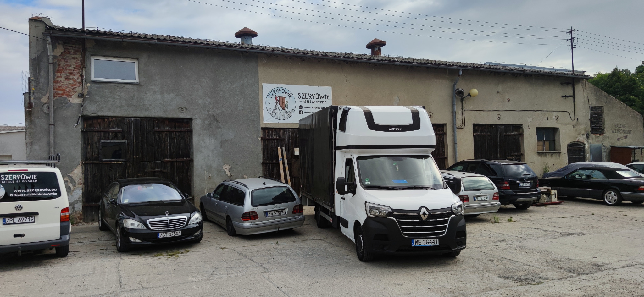 Biały bus dostawczy marki Renault z czarną plandeką, zaparkowany na betonowym placu przed budynkiem firmy meblowej 'Szerpowie', obok zaparkowane samochody osobowe.