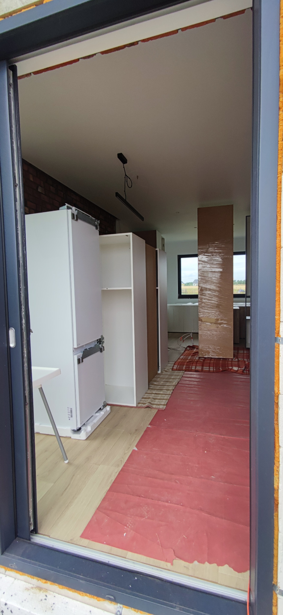 Widok przez okno na wnętrze w trakcie urządzania: białe szafy kuchenne, podłoga zabezpieczona czerwoną folią, kartonowe panele owinięte folią stretch.