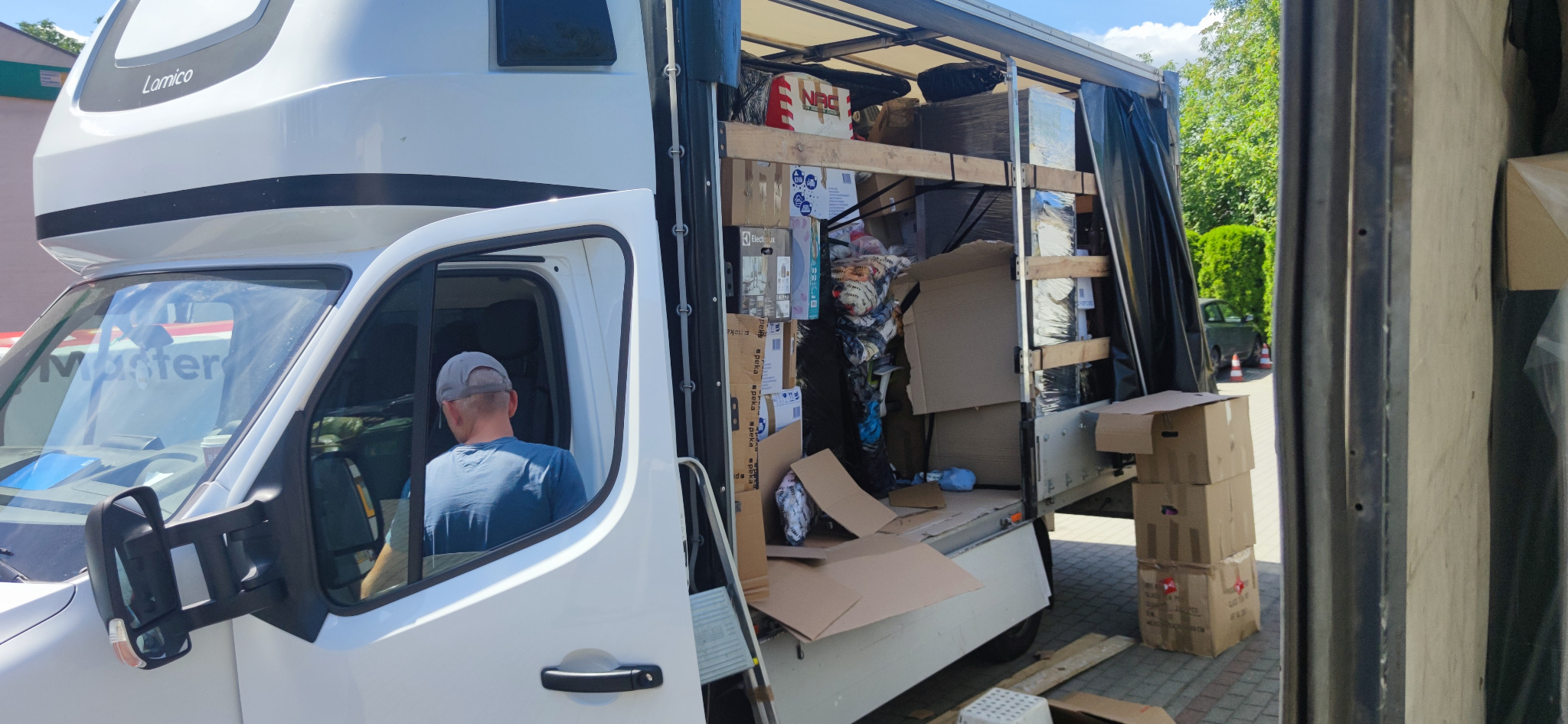 Biały samochód dostawczy z otwartą przestrzenią ładunkową, wypełnioną kartonami i różnymi przedmiotami, widok z boku na kierowcę w kabinie.