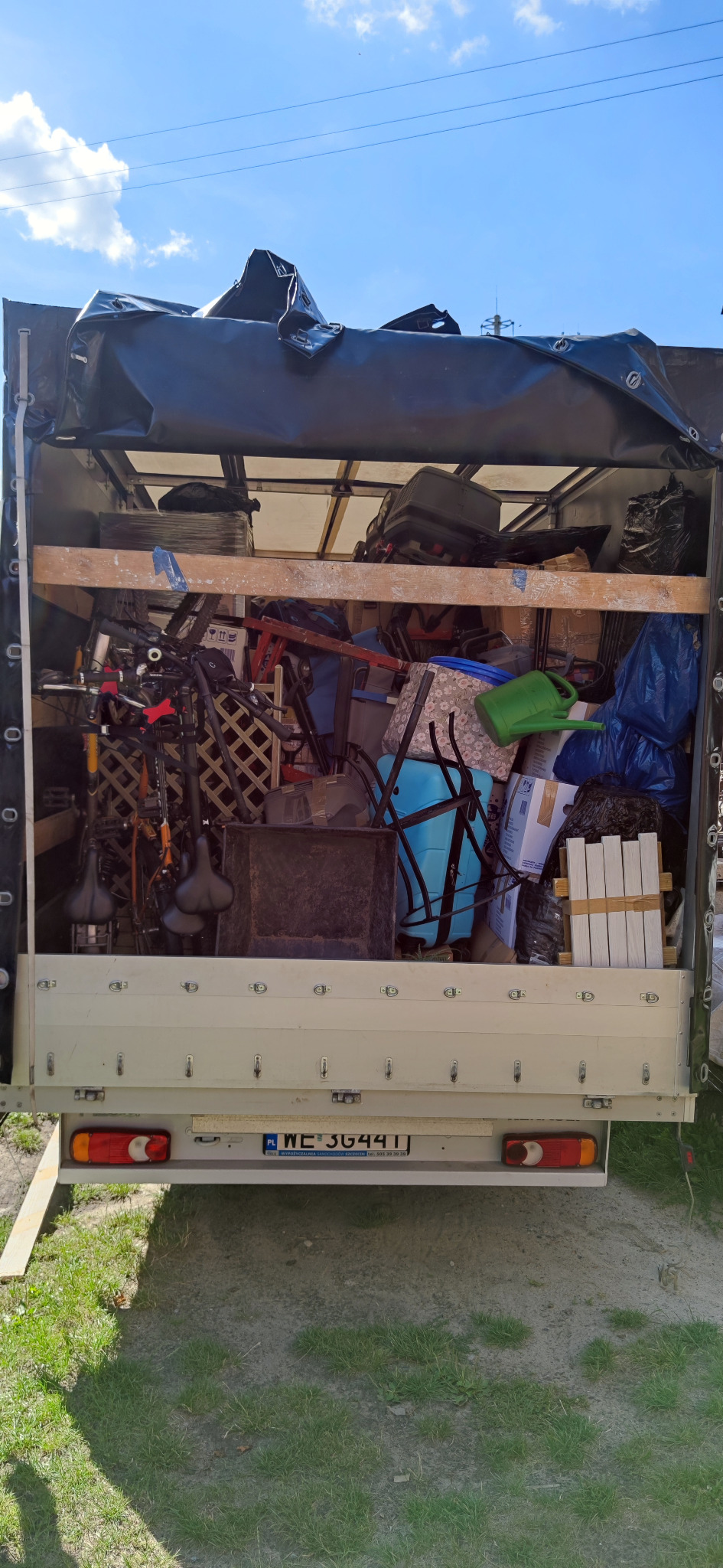 Wnętrze busa transportowego wypełnione różnorodnymi przedmiotami, w tym rowerami, meblami i kartonami, zabezpieczone czarną plandeką, widok z tyłu na tablicę rejestracyjną WE 3G4411.