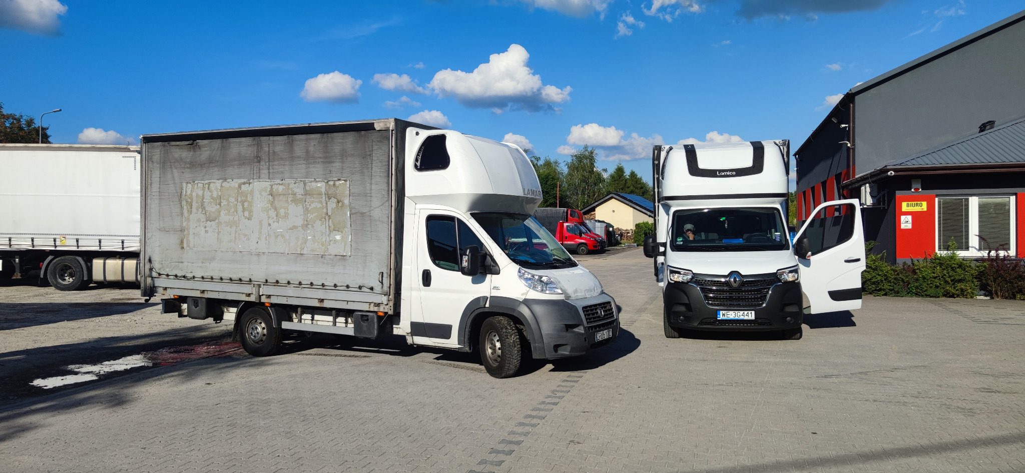 Dwa białe busy transportowe zaparkowane na placu przed budynkiem biurowym w słoneczny dzień. Jeden z busów ma otwarte drzwi kierowcy.