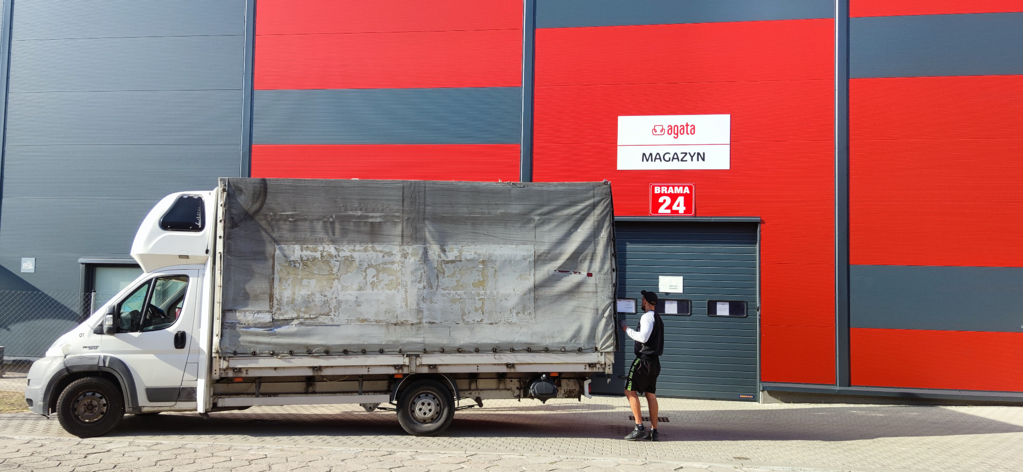 Biały samochód dostawczy z plandeką zaparkowany przed magazynem 'Agata', pracownik w czapce sprawdza dokumenty przy bramie wjazdowej.