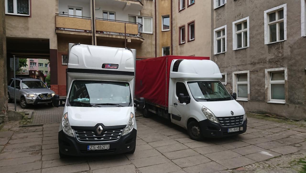 Dwa białe busy transportowe Renault Master zaparkowane na brukowanym dziedzińcu przed budynkiem mieszkalnym, jeden z czerwoną plandeką, a w tle szary samochód osobowy w przejeździe.