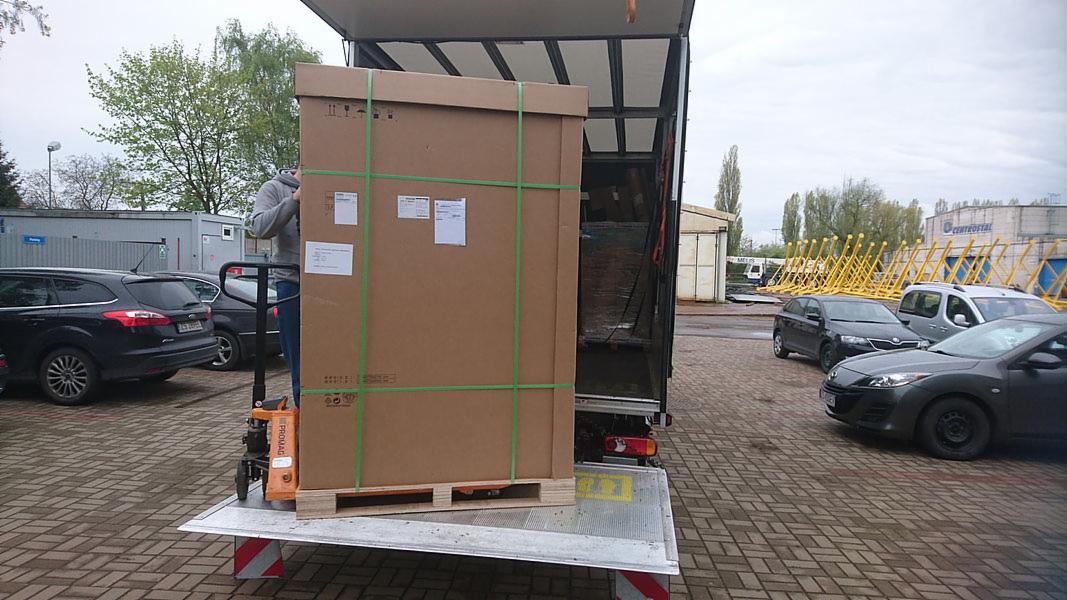 Duży karton zabezpieczony zielonymi pasami na palecie, wyładowywany z windy załadunkowej samochodu dostawczego za pomocą wózka paletowego PROMAG, na tle parkingu z samochodami osobowymi.
