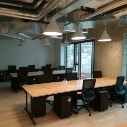 EKOCERT SPÓŁKA Z OGRANICZONĄ ODPOWIEDZIALNOŚCIĄ - Nowoczesne biuro z odsłoniętymi instalacjami wentylacyjnymi pod sufitem, jasnymi drewnianymi biurkami i czarnymi krzesłami, widok na plac budowy za oknem.
