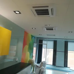 Dwa białe, kasetonowe klimatyzatory sufitowe w nowoczesnym biurze z kolorową ścianą i szklanymi ścianami działowymi. Krzesła w folii ochronnej czekają na montaż.