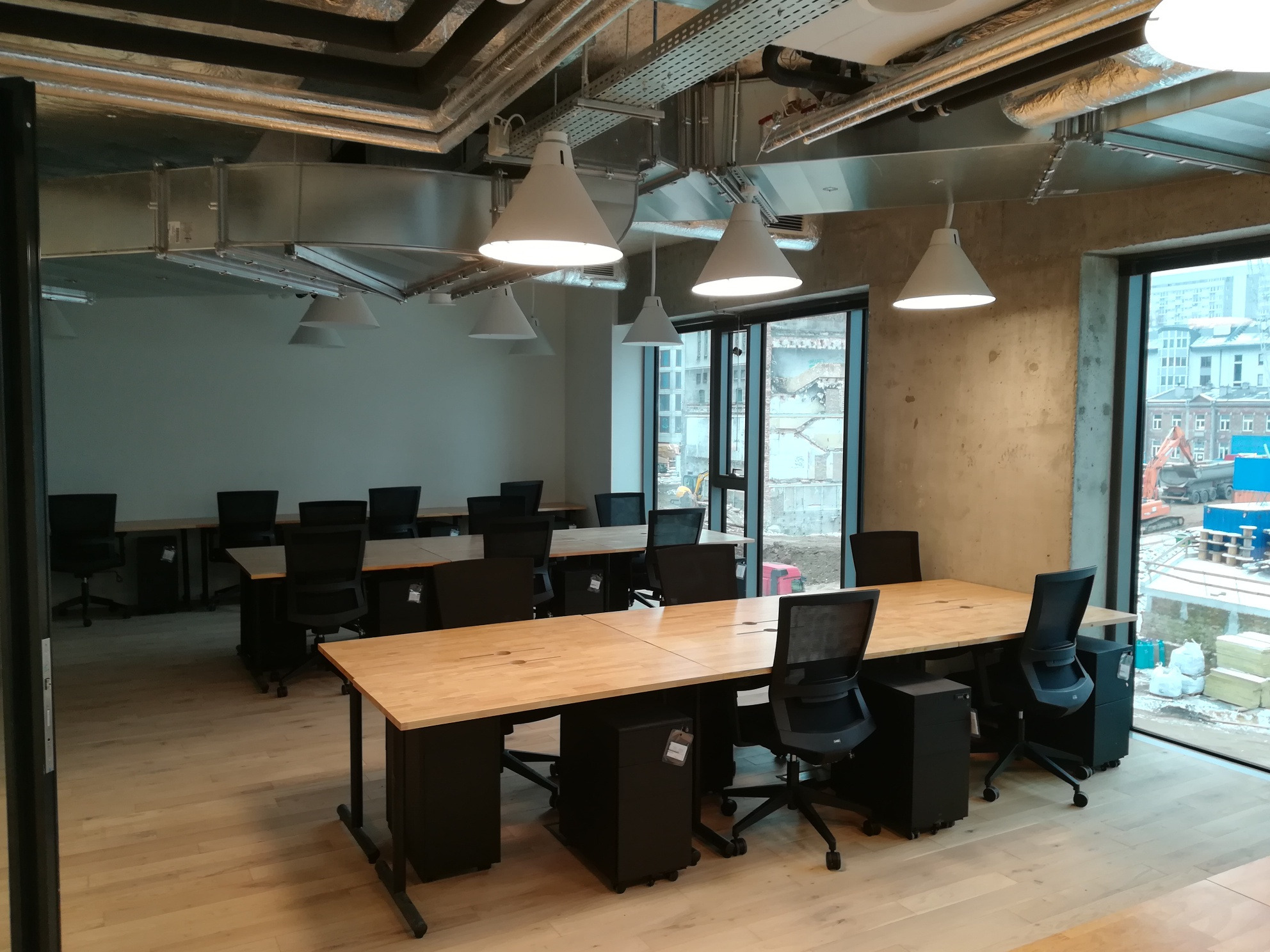 Nowoczesne biuro z odsłoniętymi instalacjami wentylacyjnymi pod sufitem, jasnymi drewnianymi biurkami i czarnymi krzesłami, widok na plac budowy za oknem.