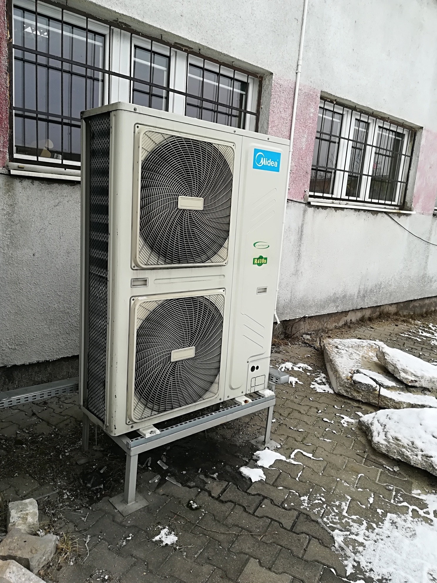 Zewnętrzna jednostka klimatyzacji Midea, dwuwentylatorowa, zamontowana na metalowym stelażu przed budynkiem z zakratowanymi oknami i resztkami śniegu na chodniku.