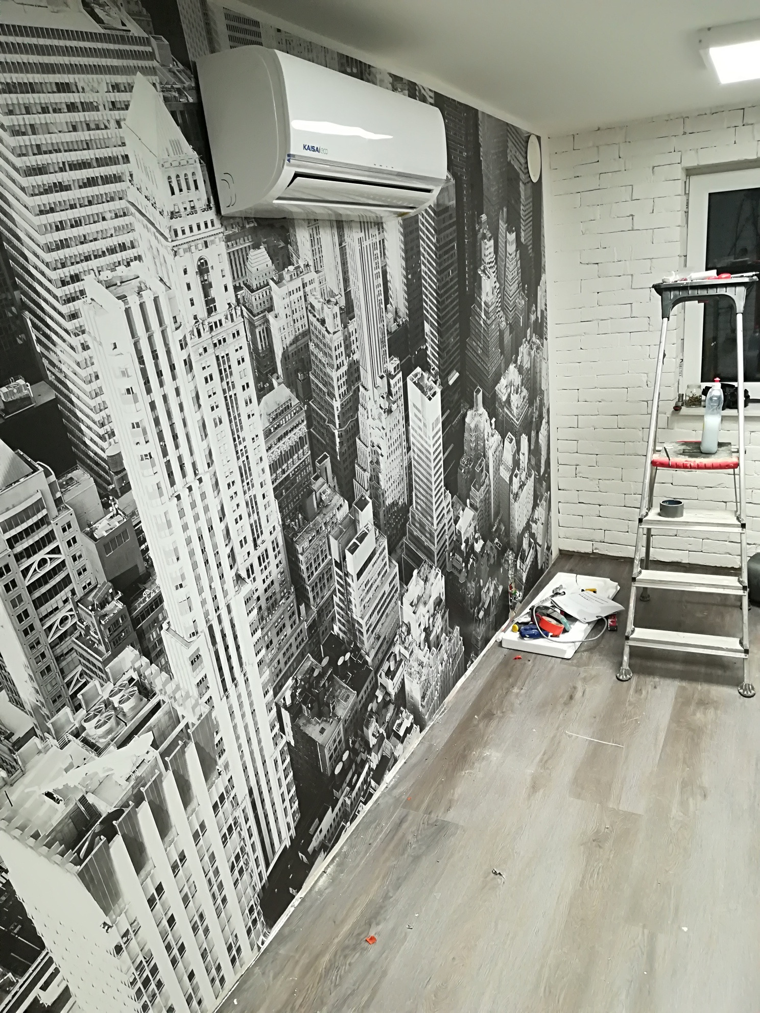 Biała jednostka klimatyzacyjna marki Kaisai zamontowana na ścianie z fototapetą przedstawiającą panoramę miasta, obok biała ściana z cegły i drabina aluminiowa.