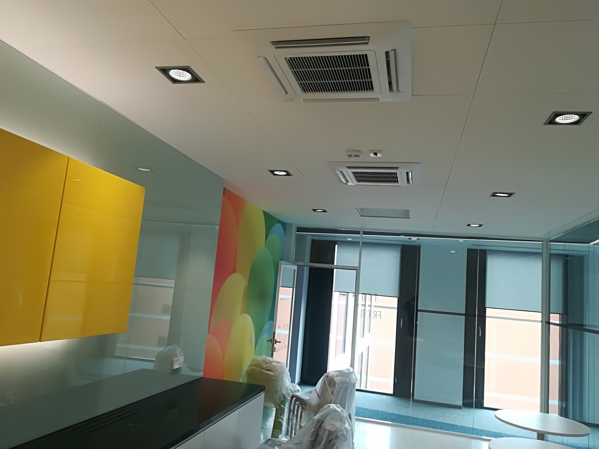 Dwa białe, kasetonowe klimatyzatory sufitowe w nowoczesnym biurze z kolorową ścianą i szklanymi ścianami działowymi. Krzesła w folii ochronnej czekają na montaż.