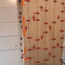 Układanie płytek imitujących drewno na ścianie, z użyciem systemu poziomowania płytek z czerwonymi klipsami i pomarańczowymi klinami, widoczny fragment wentylacji.