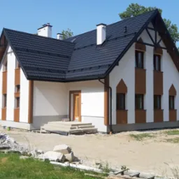 Nowo wybudowany dom jednorodzinny z białą elewacją i brązowymi akcentami, czarnym dachem i widocznymi śladami prac budowlanych wokół budynku.