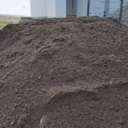 Kupa ciemnej ziemi usypana przed budynkiem i metalowym ogrodzeniem, widoczne ślady po szpadlu.