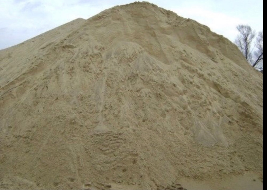 Góra piasku o drobnej strukturze, z widocznymi śladami osypywania się, na tle pochmurnego nieba i fragmentu drzewa.
