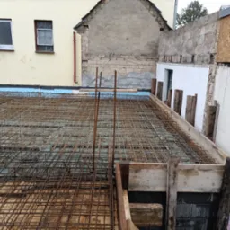 Zbrojenie fundamentu z widoczną siatką stalową i szalunkiem drewnianym. W tle fragment budynku z oknami i murem. Widok z góry na przygotowany teren pod wylewkę.