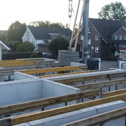 Przychodaj Konrad - Fundamenty domu jednorodzinnego w trakcie budowy, widoczne szalunki i bloczki fundamentowe, w tle zabudowa mieszkalna i dźwig budowlany.