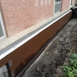 Izolacja fundamentów budynku z cegły, zabezpieczona brązową powłoką hydroizolacyjną, widoczne okno i fragment terenu.