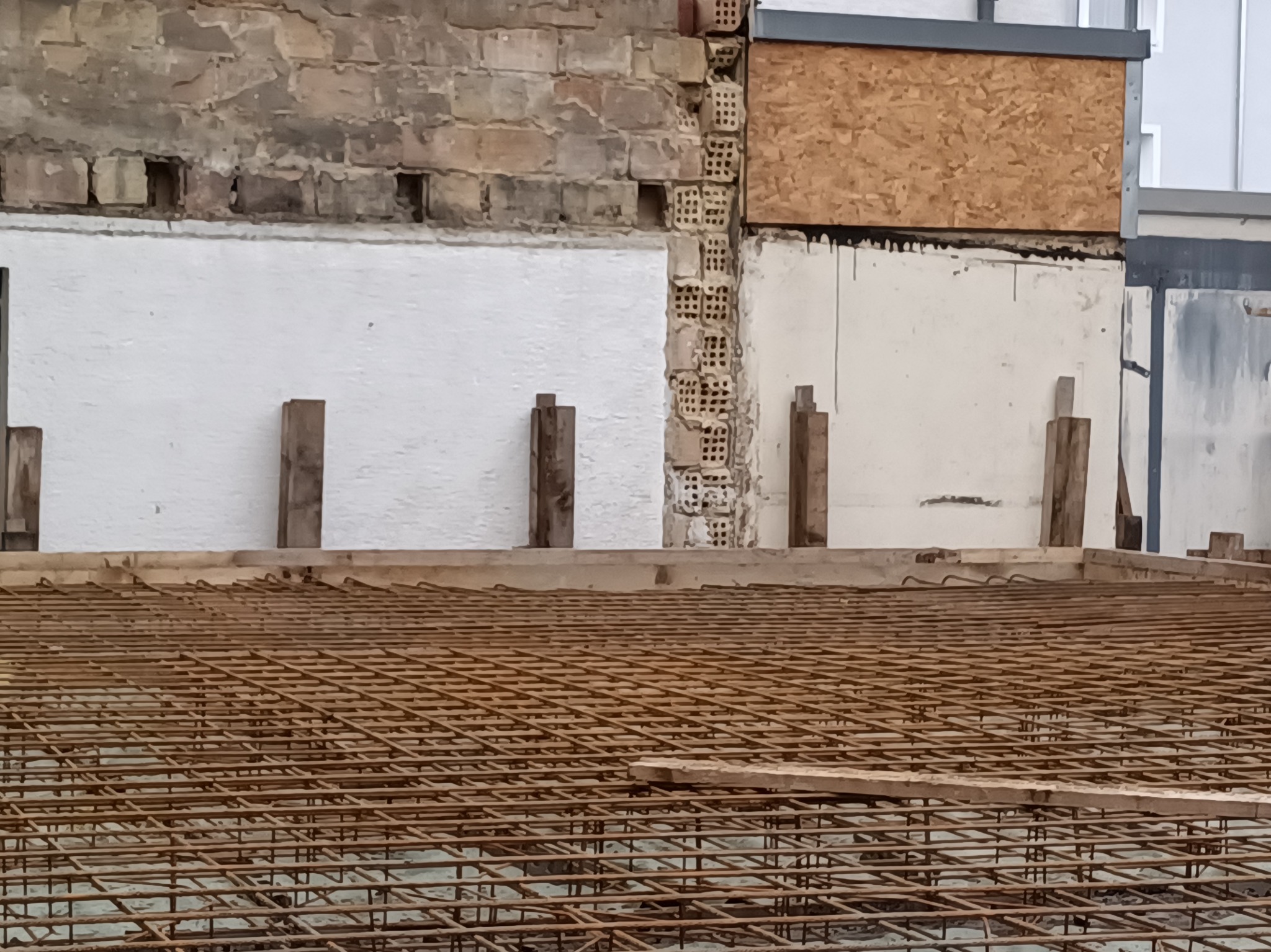 Zbrojenie fundamentów pod budynek z odsłoniętym fragmentem ceglanej ściany i białym tynkiem. Widoczne drewniane podpory i elementy konstrukcyjne.