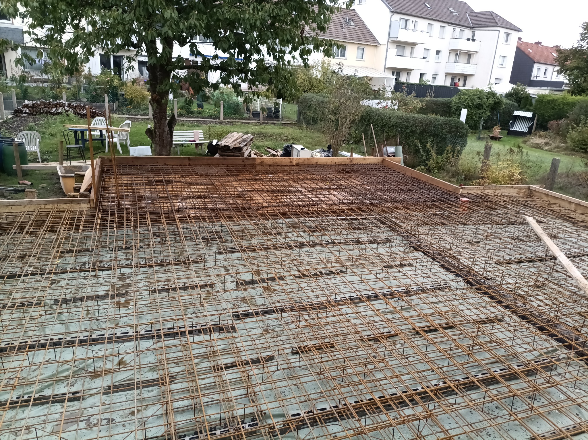 Zbrojenie fundamentu domu jednorodzinnego, widok z góry na siatkę z prętów stalowych, szalunek drewniany. W tle ogród i zabudowa mieszkalna.