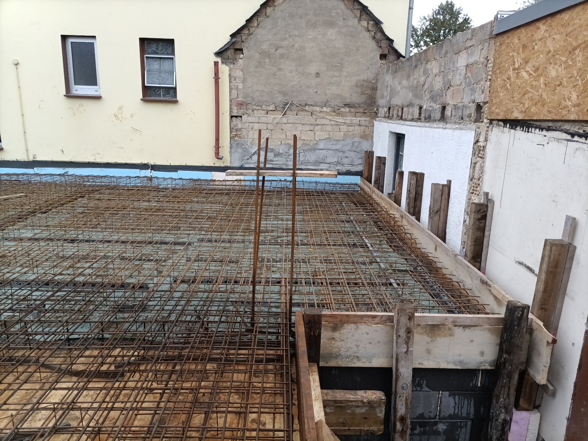 Zbrojenie fundamentu z widoczną siatką stalową i szalunkiem drewnianym. W tle fragment budynku z oknami i murem. Widok z góry na przygotowany teren pod wylewkę.