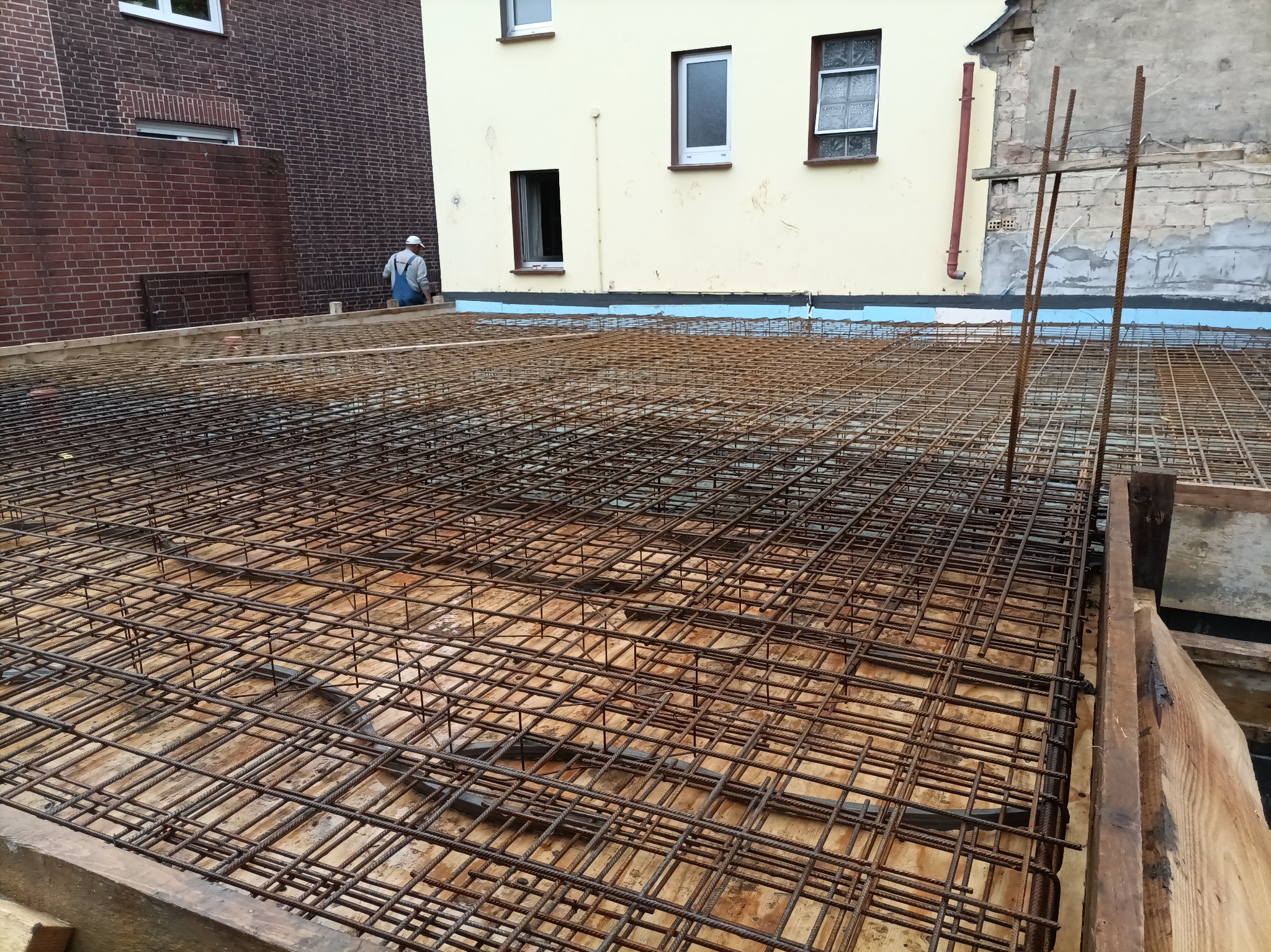 Siatka zbrojeniowa na deskowaniu fundamentu, zabezpieczona izolacją, z widocznym pracownikiem w tle i budynkami w różnym stanie wykończenia.