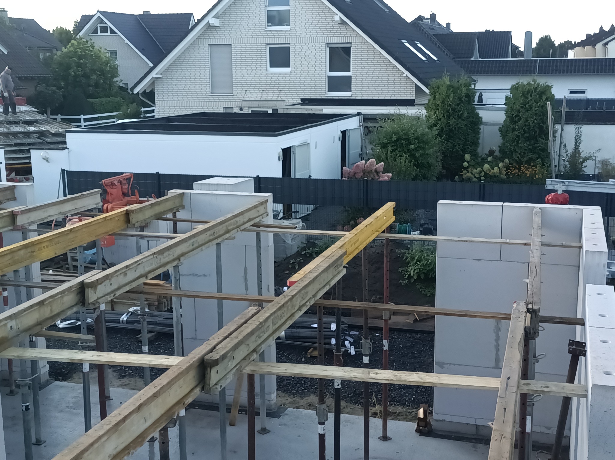 Budowa domu: wznoszenie ścian z bloczków, podparte drewnianymi belkami i metalowymi stemplami. W tle zabudowa jednorodzinna i elementy ogrodu.