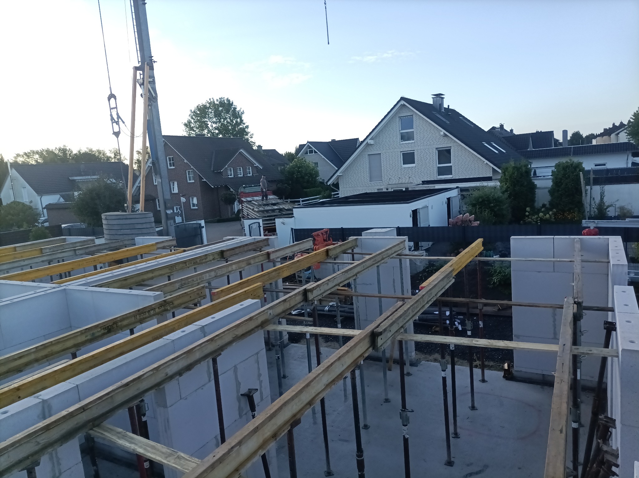 Budowa domu: widok z góry na fundamenty z białych bloczków, drewniane belki wzmacniające, i podpory. W tle domy mieszkalne i żuraw budowlany.