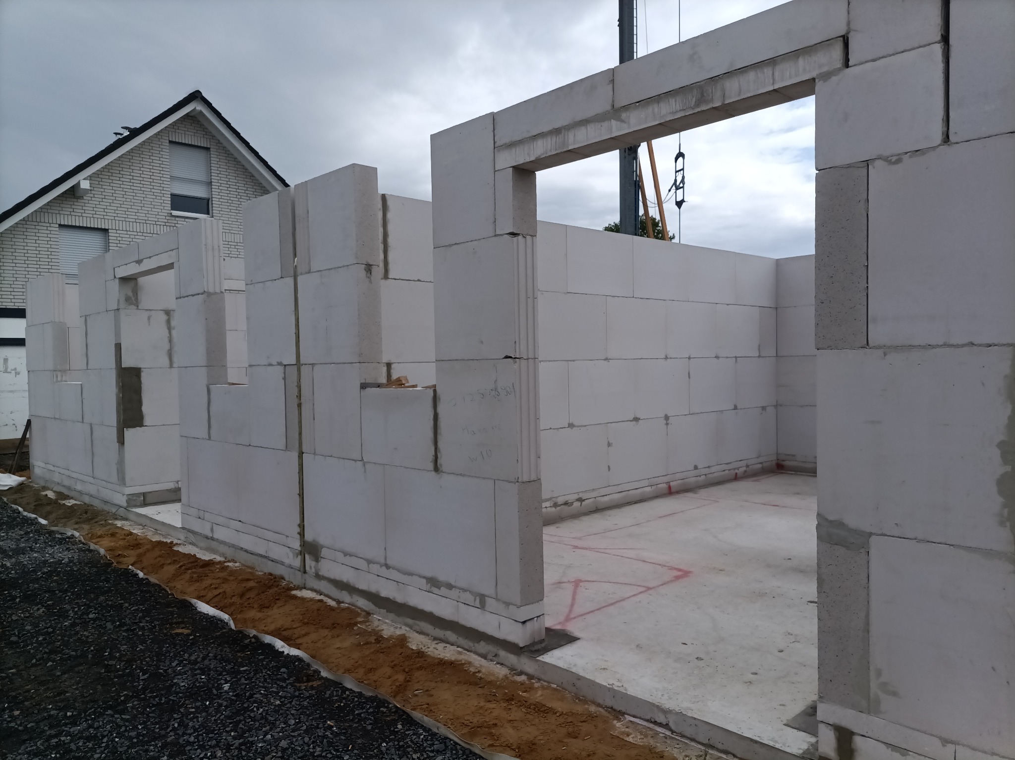 Wznoszenie ścian domu z betonu komórkowego Ytong. Widoczne fragmenty ścian z otworami okiennymi i drzwiowymi. Na drugim planie dom w trakcie budowy i żuraw.