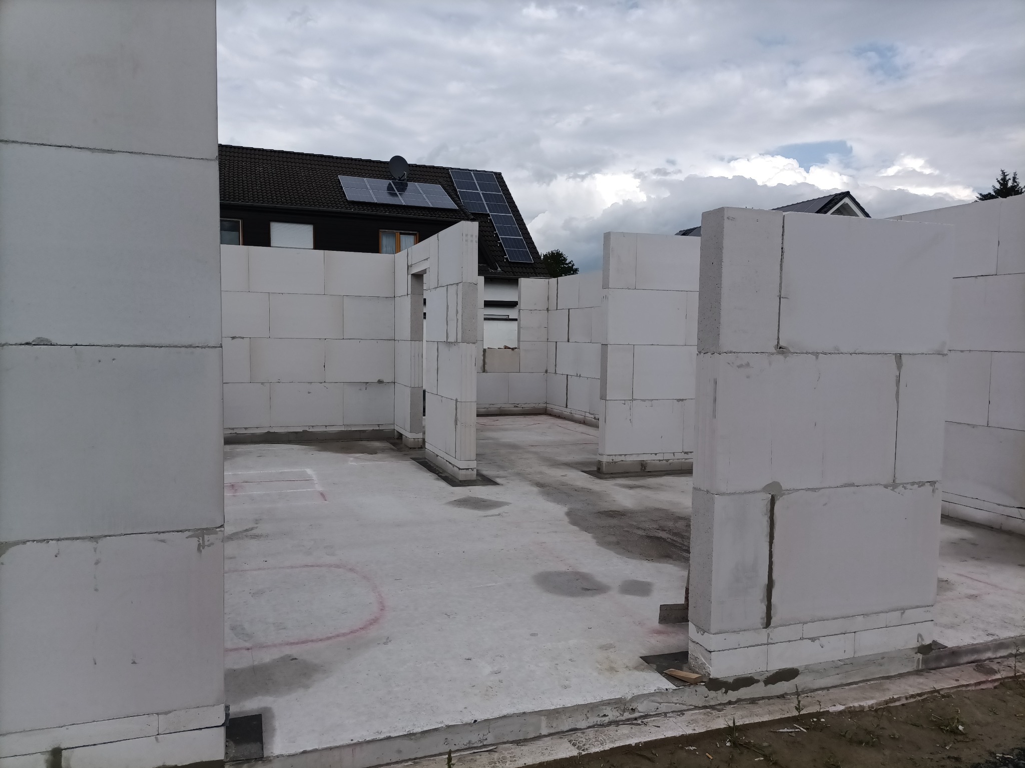Wnętrze w trakcie budowy domu z białych bloczków, widoczne ściany nośne i fundamenty. Na dachu budynku w tle panele słoneczne.