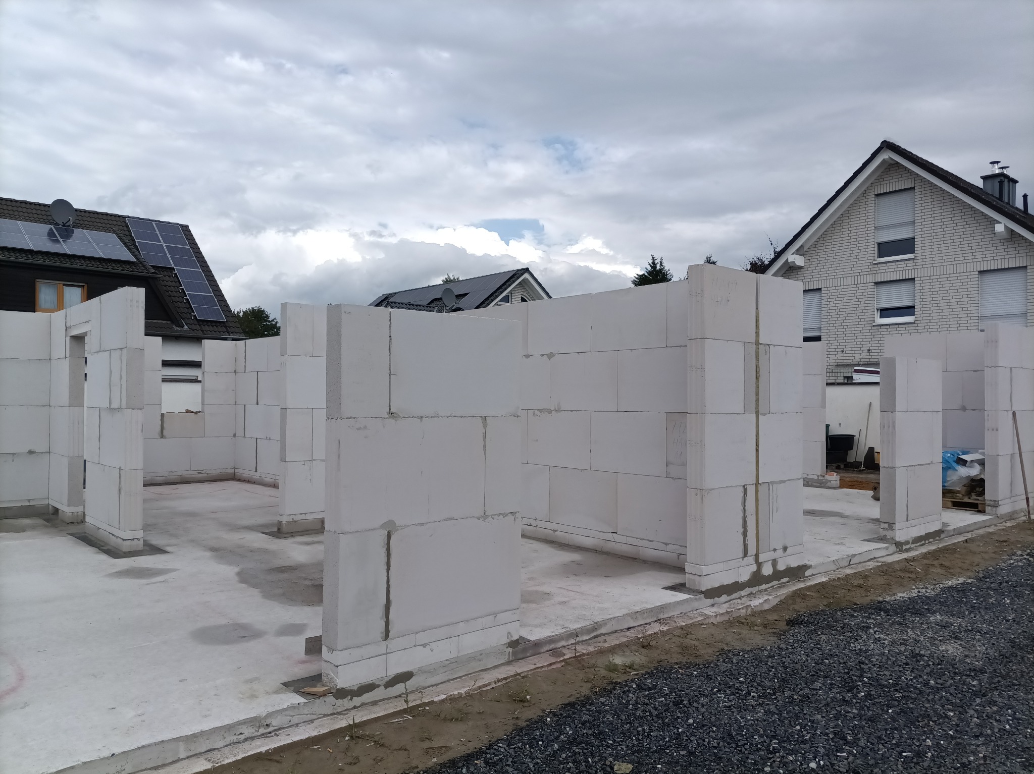 Wnętrze domu w trakcie budowy z białych bloczków, widoczne fragmenty ścian i słupów. W tle dom z panelami słonecznymi i pochmurne niebo.