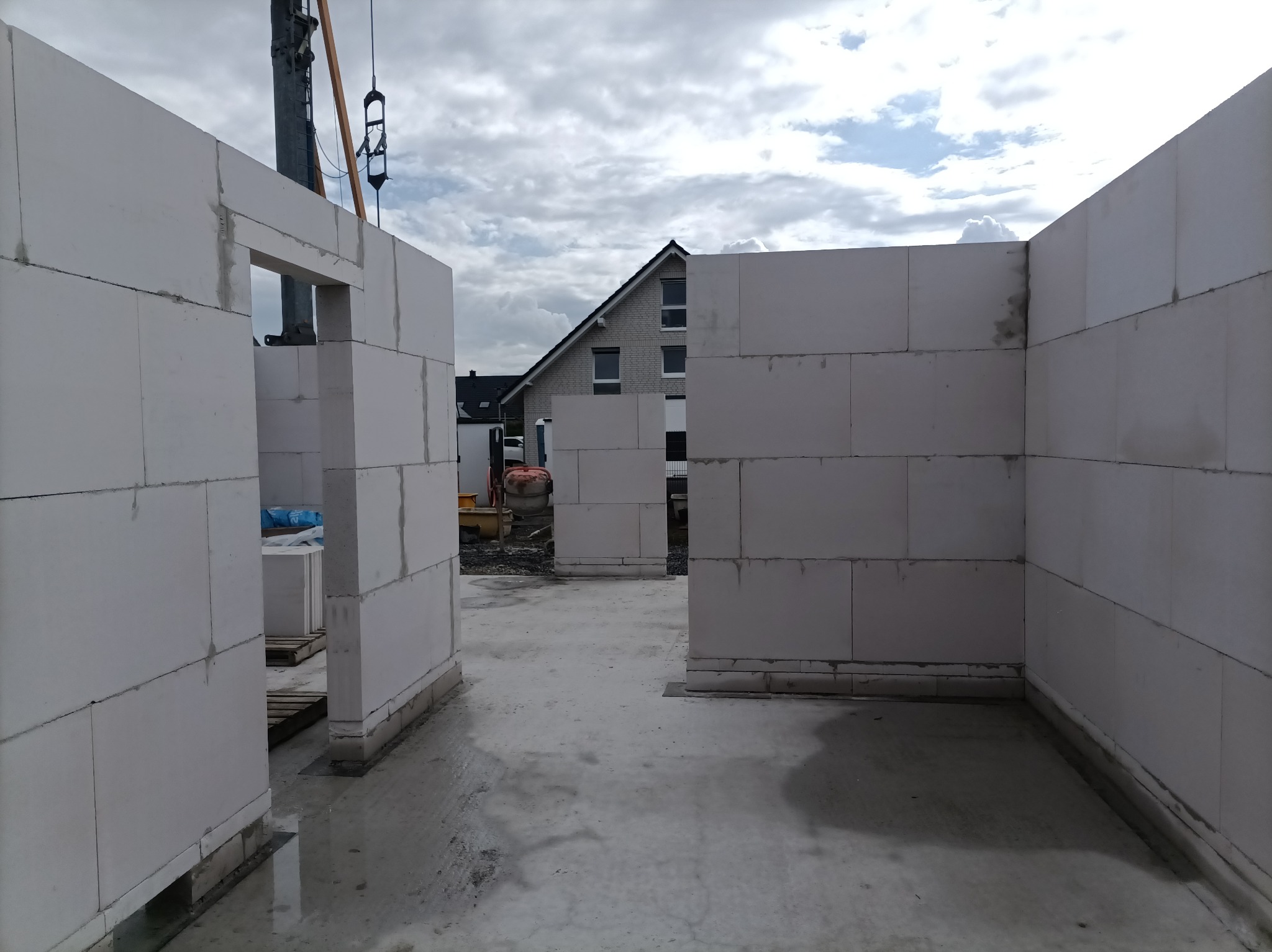 Wnętrze surowego domu w trakcie budowy z białych bloczków, z widocznym dźwigiem budowlanym i fragmentem zabudowy jednorodzinnej w tle.
