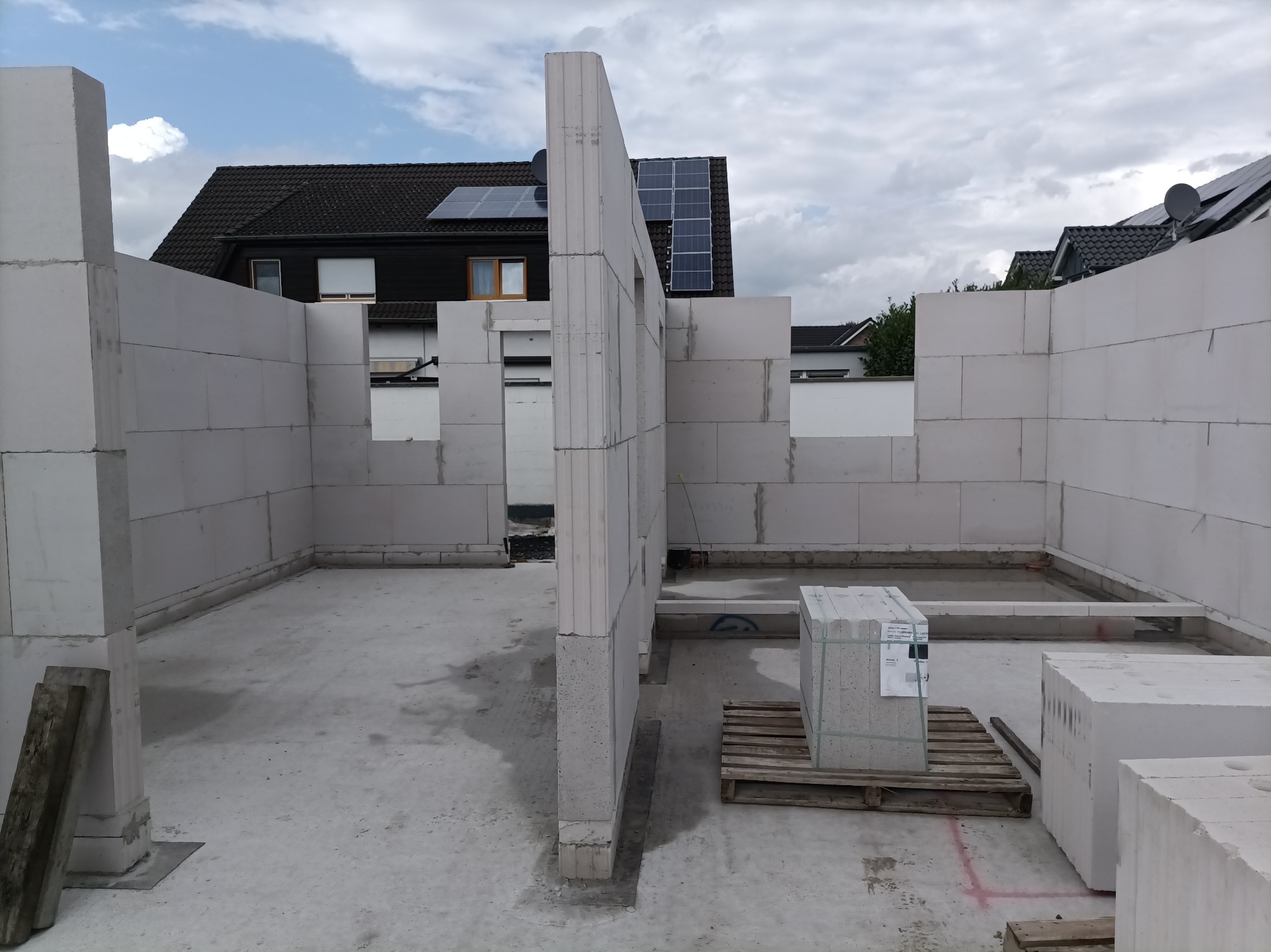 Wnętrze domu w trakcie budowy z białych bloczków, widoczne ściany nośne, okna, paleta bloczków. W tle dom z panelami słonecznymi. Pochmurne niebo.