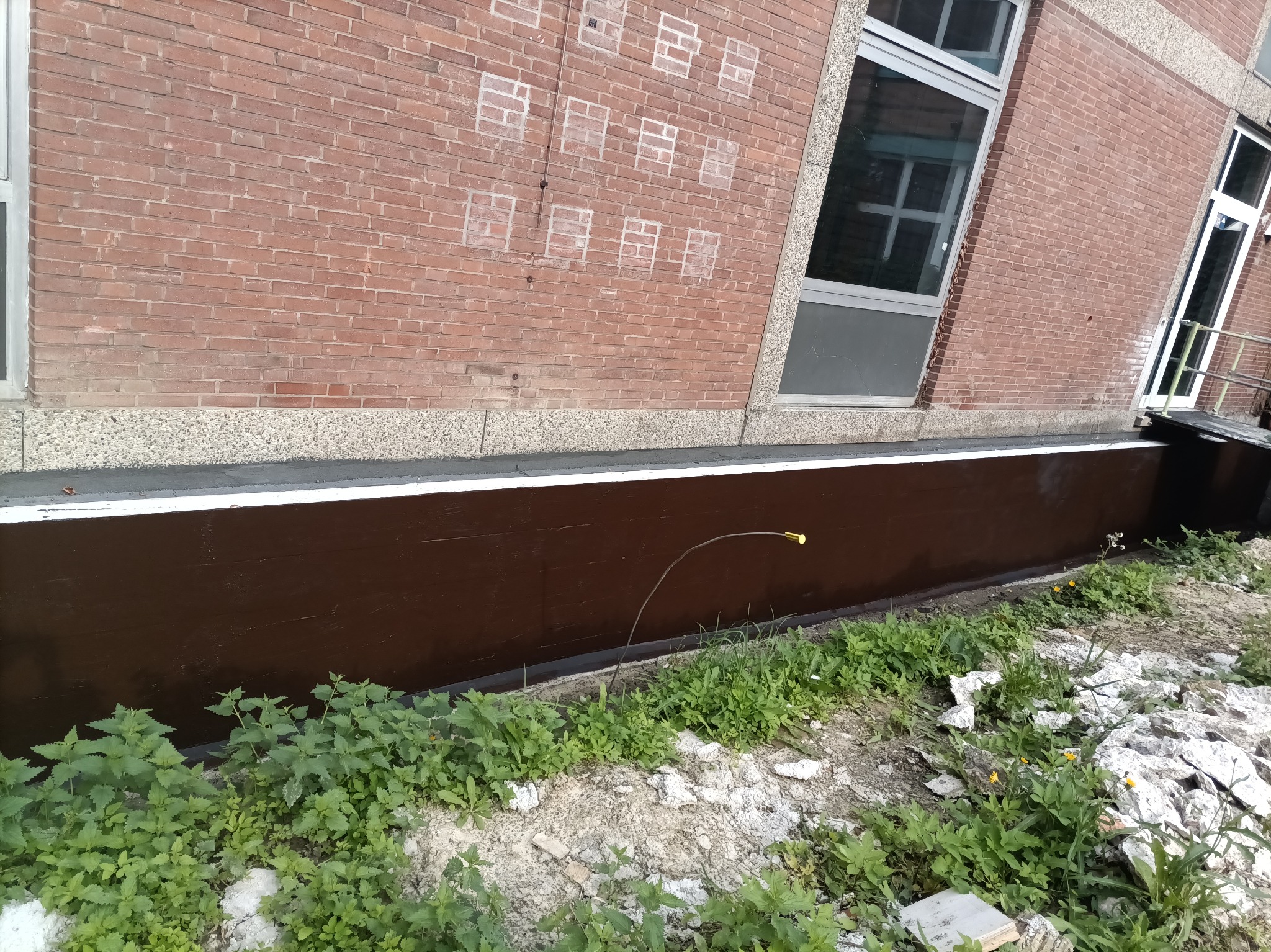 Boczny widok izolacji fundamentu budynku z cegły, z widocznymi oknami i roślinnością u podstawy. Brązowa powłoka hydroizolacyjna na fundamencie.