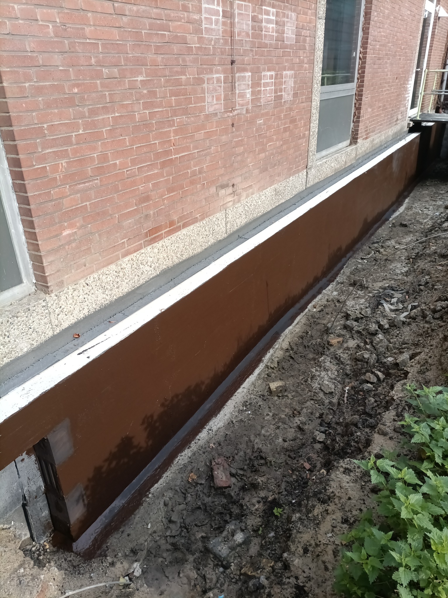 Izolacja fundamentów budynku z cegły, zabezpieczona brązową powłoką hydroizolacyjną, widoczne okno i fragment terenu.